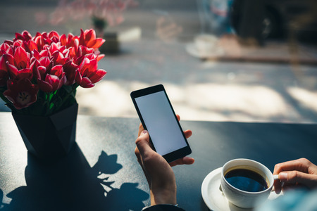 Close-up of woman's hand holding mobile phone with blank screen next to cup of coffee and bouquet of flowers at table in cafe near window.の写真素材