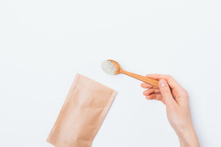 Cosmetic clay in wooden spoon on white background, flat lay. Woman's hand gets ingredient for face...