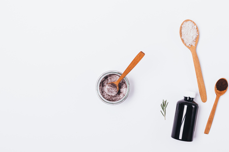 Flat lay composition jar of homemade skin scrub near ingredients: bottle of cosmetic oil, sea salt, ground coffee and sprig of rosemary on white background with copy space.の写真素材