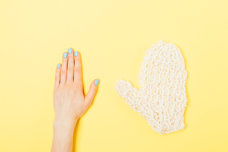 Flat lay concept of body care bath accessories on bright yellow background. Minimal composition of woman's hand with blue manicure next to coarse mitten washcloth.の写真素材