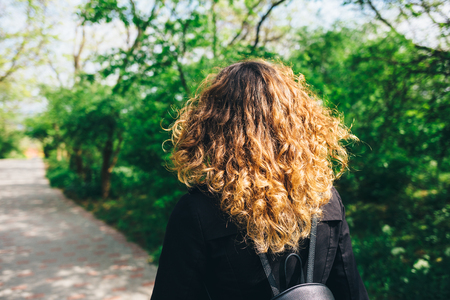 Back view young woman with thick natural brown curly hair walking along green park on sunny spring day.の写真素材
