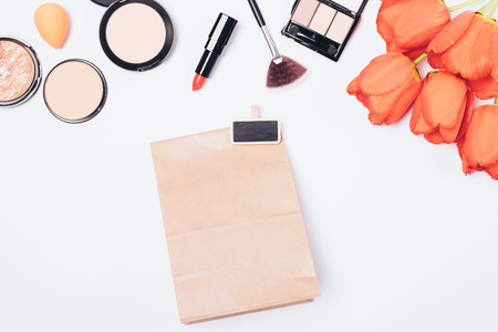 Flat lay makeup background of women's lipsticks, brushes and powders with bouquet of flowers next to gift brown paper bag, top view on white table.の写真素材