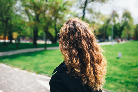Close-up of young woman with curly hair walking in park on sunny spring day, back view.の写真素材