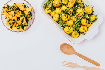 Vegan dish baked new potatoes with green beans next to salad of fresh yellow tomatoes and parsley, top view on white background.の写真素材
