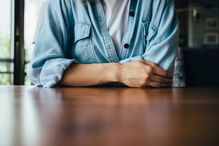 Folded hands of young woman wearing blue casual shirt and white t-shirt sitting at empty wooden...