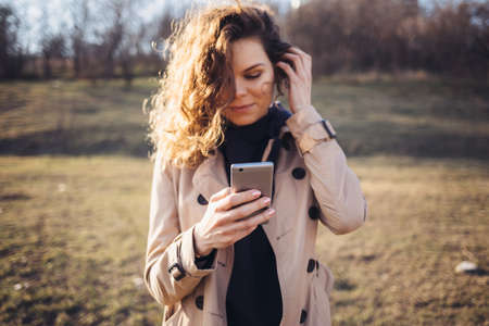 Smiling young woman with long curly hair wearing a beige coat stands outdoors in a grass field on an autumn day holding texting on her mobile phone.の写真素材
