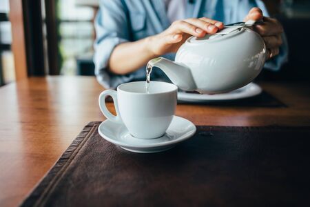Female's hands pour tea from teapot into cup, close-up. Young woman prepares hot drink sitting at...