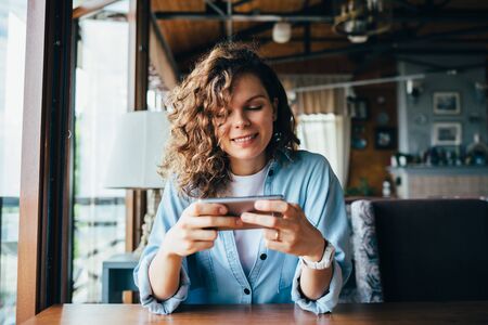 Young woman wearing blue shirt and white t-shirt smiles while texting on smart phone sitting at window at restaurant table in daytime.の写真素材