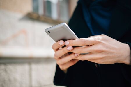 Young woman using smart phone standing outside in city at daytime. Female's hands with neutral manicure holding mobile device, close-up.の写真素材