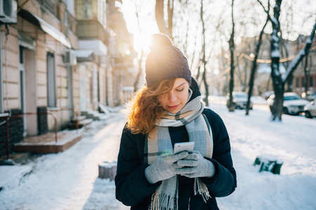 Young woman wearing warm scarf and knitted hat smiles and using mobile phone writing text message outdoors on sunny winter day in city.の写真素材