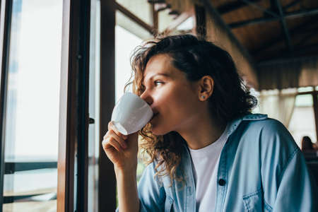 Young woman with long curly hair wearing denim shirt drinking coffee looks out the window sitting in restaurant in the daytime.の写真素材