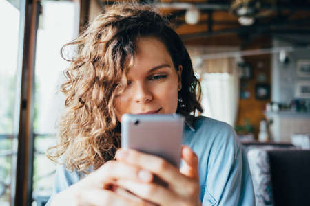 Close-up portrait of young woman texting on smart phone sitting in restaurant by the window in daytime and smiling looking at screen.の写真素材