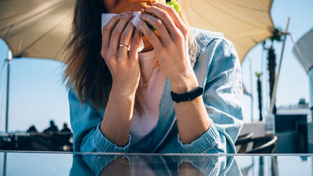 Close-up of young woman eating hamburger sitting in outdoor cafe at sunny day.の写真素材