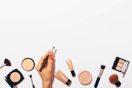 Bottom frame composition of female's hand holding professional makeup brush among beauty products, powders, and concealers on white background with copy space.の写真素材