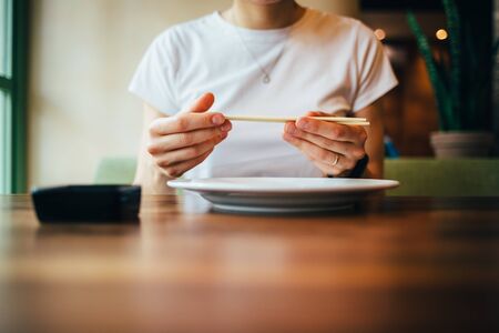 Close-up of female hands holding chopsticks. Young woman is preparing to eat sushi sitting at table in restaurant.の写真素材