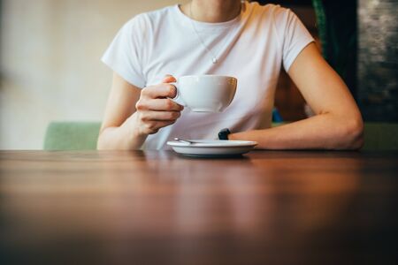Close-up of female's hands holding cup of coffee. Woman drinking cappuccino sitting at table in...