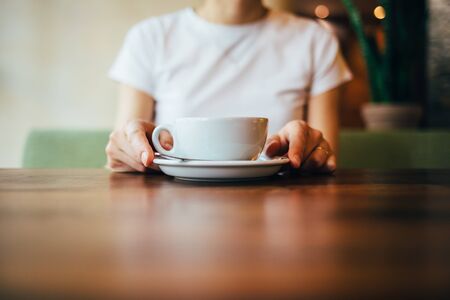 Close-up of female's hands holding saucer with cup of coffee. Young woman preparing to drink cappuccino at table in cafe at daytime.の写真素材
