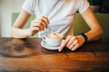Female's hands stir with spoon cappuccino with foam, close-up. Young woman drinking coffee sitting at wooden table in cafe in daytime.の写真素材