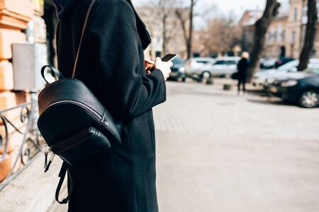Side view young woman wearing elegant black coat and backpack using mobile phone standing on street in old European city near road with cars in daytime, copy space.の写真素材