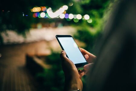 Close-up of young woman holding smart phone with blank white screen walking down the street at summer night, festive colored lights on the background.の写真素材