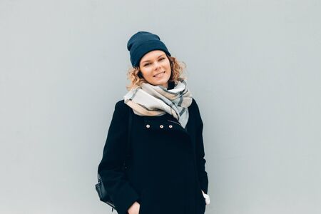 Happy female outdoors near wall in winter wearing coat, scarf, hat and backpack. Young woman...