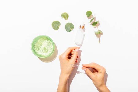 Female's hands applying cosmetic moisturizing aloe gel next to bottle of serum and branch with eucalyptus leaves on white background, flat lay.の写真素材