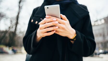 Close-up of a woman in a black coat looks at the screen of a mobile phone in the cityの写真素材