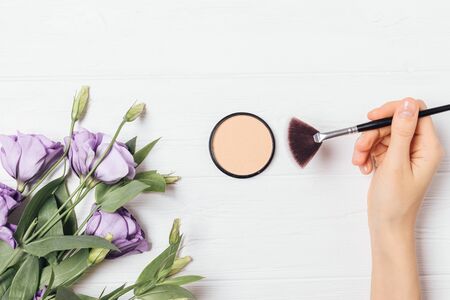Female hand holding makeup brush using pressed powder on white background with purple lisianthus flowers, flat lay composition.の写真素材