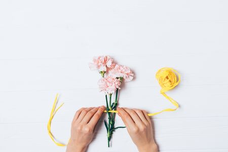 Flat lay woman's hands decorating bouquet of fresh pale pink flowers with yellow ribbon on white background.の写真素材