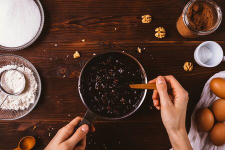 Female's hands mix melted chocolate with nuts and dried cherries in saucepan among ingredients for baking delicious homemade brownie cake, top view.の写真素材
