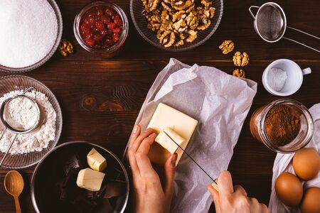 Woman's hands cut butter among ingredients for baking homemade brownie, flour, eggs, cocoa, chocolate and delicious cherries and nuts additives on wooden table, top view.の写真素材