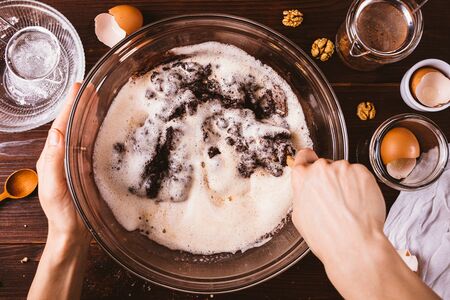 Woman's hands mix whipped eggs with melted chocolate and nuts for delicious brownie cake in large bowl with spoon on dark wooden table next to ingredients and cooking utensils, top view.の写真素材