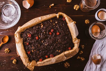 Cooked brownie cake in baking dish with parchment among ingredients on dark wooden table, flat lay.の写真素材