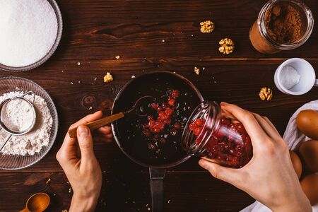 Top view female hands adding dried cherries to melted chocolate with chopped nuts to make dough for...