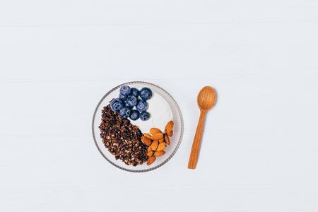 Granola with yogurt, blueberries and almonds near wooden spoon on white table, top view.の写真素材