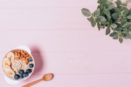 Flat lay healthy breakfast bowl of oatmeal with almond nuts, blueberries and bananas near green houseplant on pink table.の写真素材