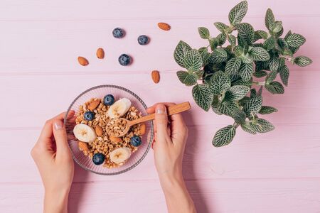 Woman's hands holding bowl of healthy granola breakfast with nuts and berries next to green houseplant on pink table, top viewの写真素材