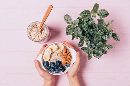 Female's hands holding bowl of delicious oatmeal with blueberries, almonds and bananas next to jar and green plant on pink table, top view.の写真素材