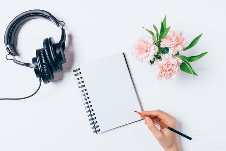 Woman's hand writing in blank notepad near headphones and bouquet of flowers on white desk, flat lay. Feminine workspace concept.の写真素材