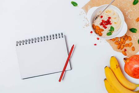 Healthy breakfast of oatmeal with nuts, pomegranate seeds and bananas next to blank notepad on white table, flat lay.の写真素材