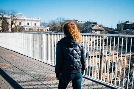 Rear view of young woman with curly hair dressed in short jacket walks over city bridge on sunny...
