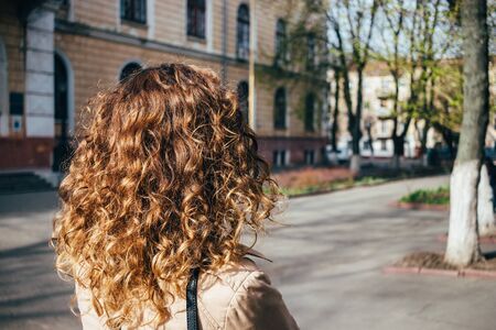 Rear view woman with natural lush wavy hairstyle walking in european city. Curly hair close-up outside on sunny spring day.の写真素材