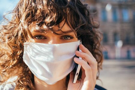 Portrait young woman wearing medical face mask calling on cell phone while standing on street in the city.の写真素材