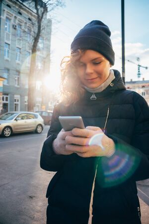 Happy young woman wearing warm clothing using mobile phone writing text message outdoors on sunny winter day in city.の写真素材