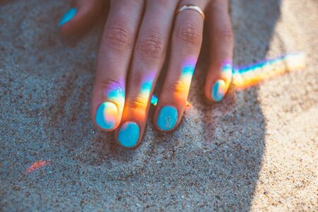 Young woman hand with blue manicure and reflection of rainbow on sand, close-up.の写真素材