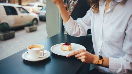 Young woman eating cheesecake drinking coffee sitting cafe near window, close-up.の写真素材