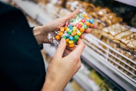 Female hands holding package of candies near shelf in confectionery department of supermarket.
