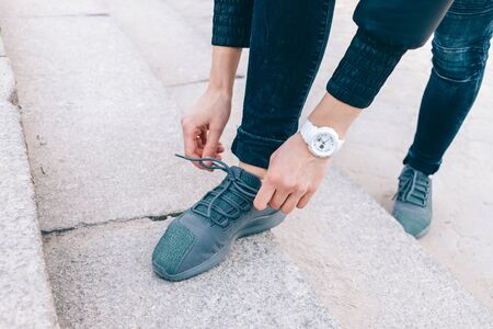 Close-up of young woman tying shoelaces on her sneakers while standing on street in city.
