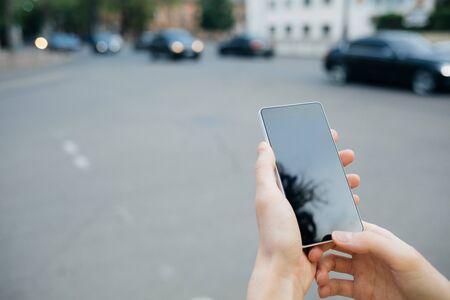 Female hands holding mobile phone with blank display near road with cars in city, close-up.の写真素材