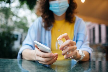 Young woman in medical face mask spraying alcohol disinfectant on her cell phone while sitting at a table in a street restaurant in the city.の写真素材